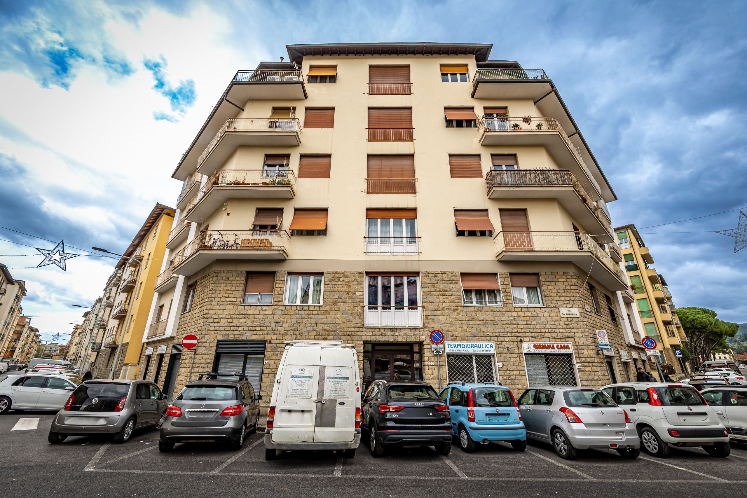 Un edificio residencial de varios pisos con balcones, entrada a nivel de calle y un estacionamiento con varios automóviles.