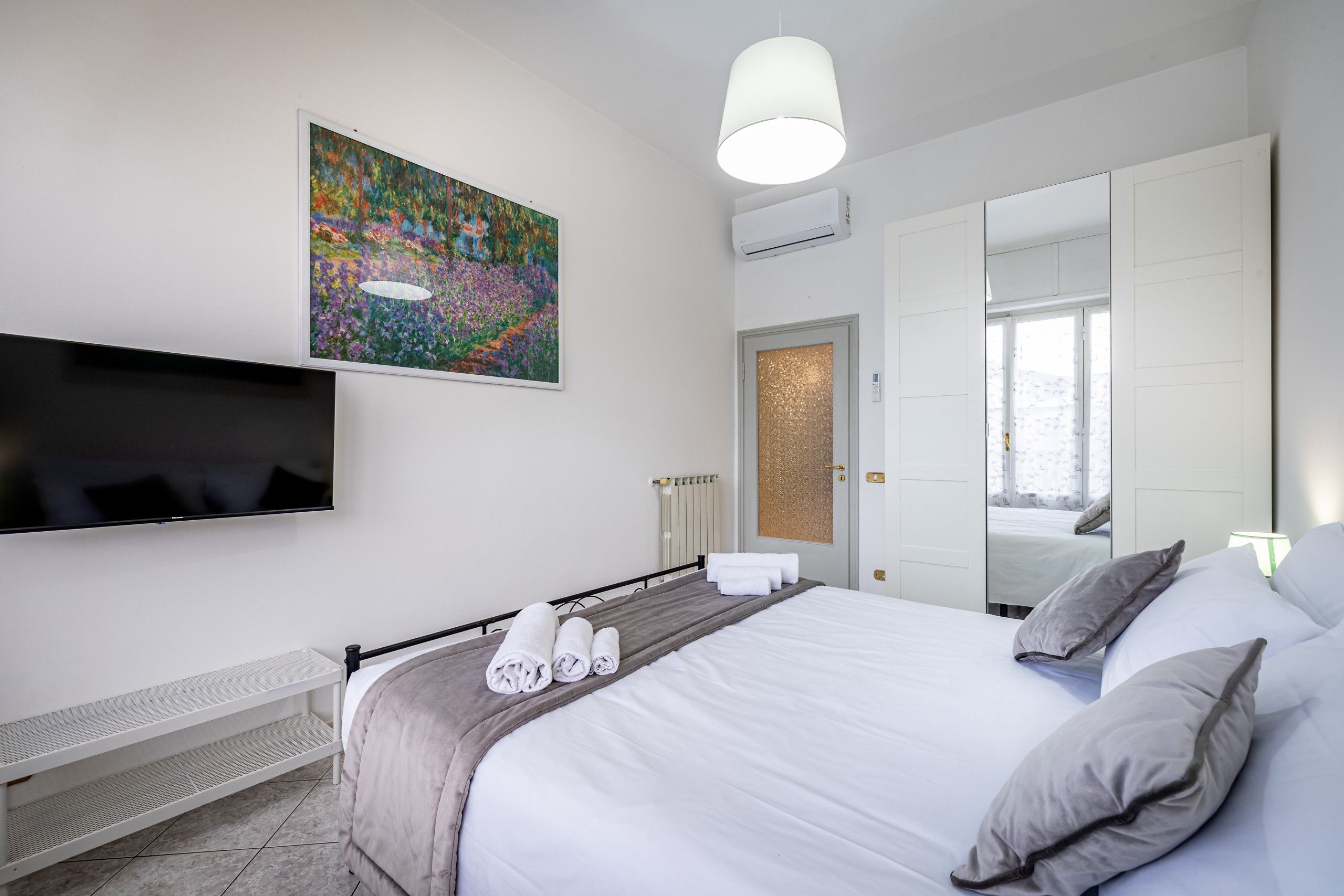Una habitación de hotel moderna con una cama grande, un televisor de pantalla plana montado en la pared, un cuadro sobre la cama y un espejo grande que refleja la habitación.