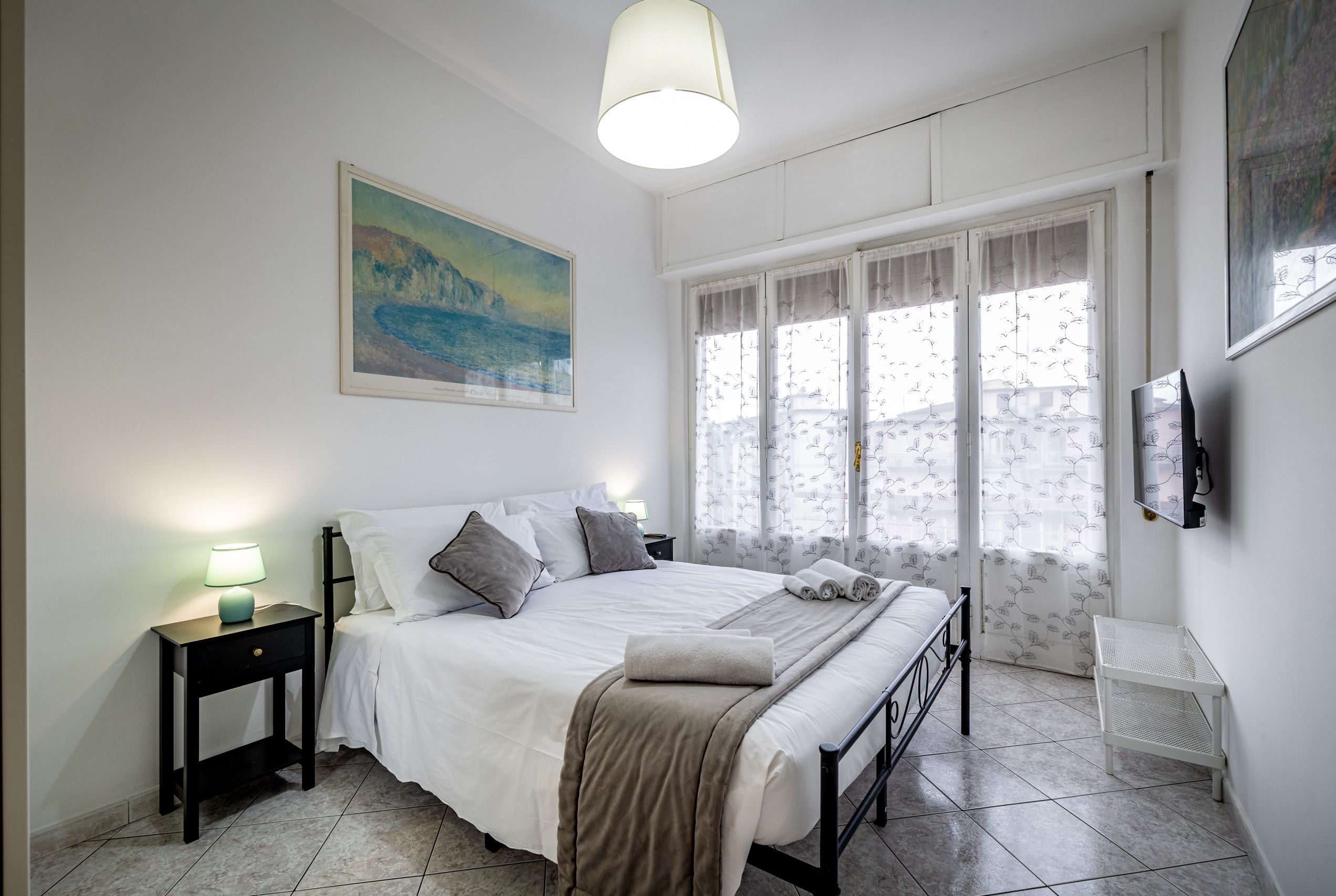 A bedroom with a bed, a nightstand, a painting, a window with curtains, and a TV on the wall.