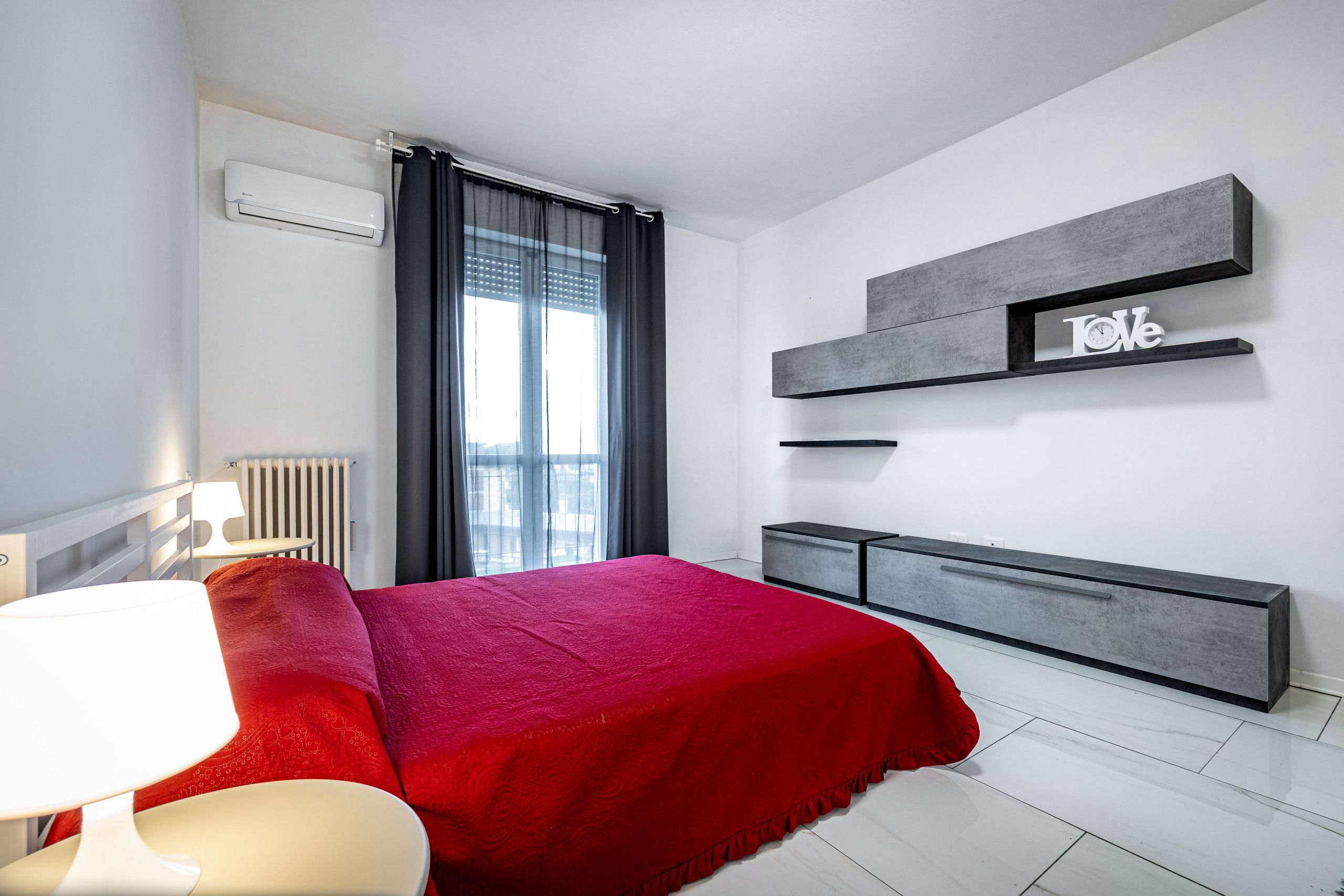 Un dormitorio moderno con cama, mesitas de noche, radiador y ventana con cortinas.
