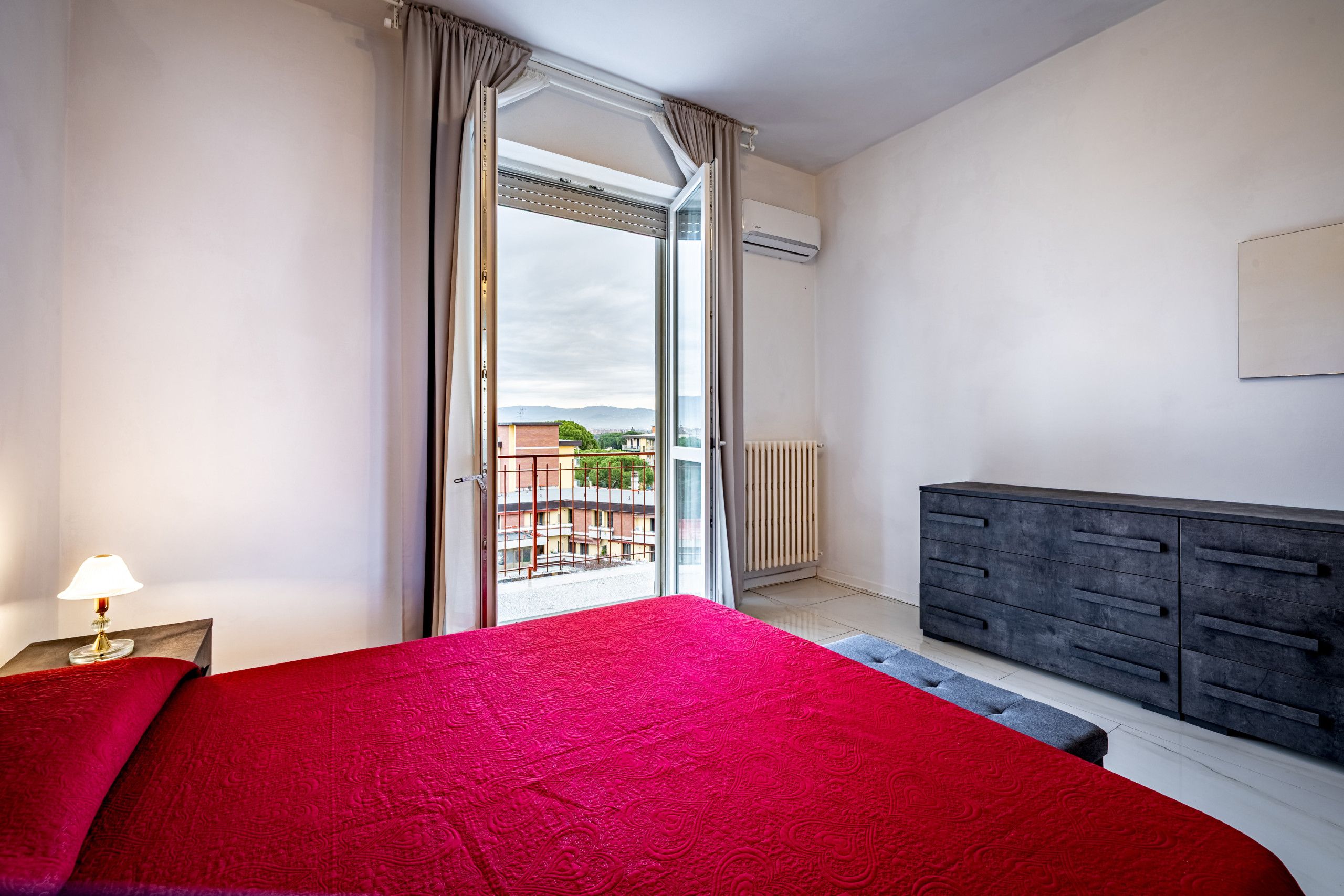 Un dormitorio con cama, cómoda, radiador y vista a edificios a través de una puerta de cristal.