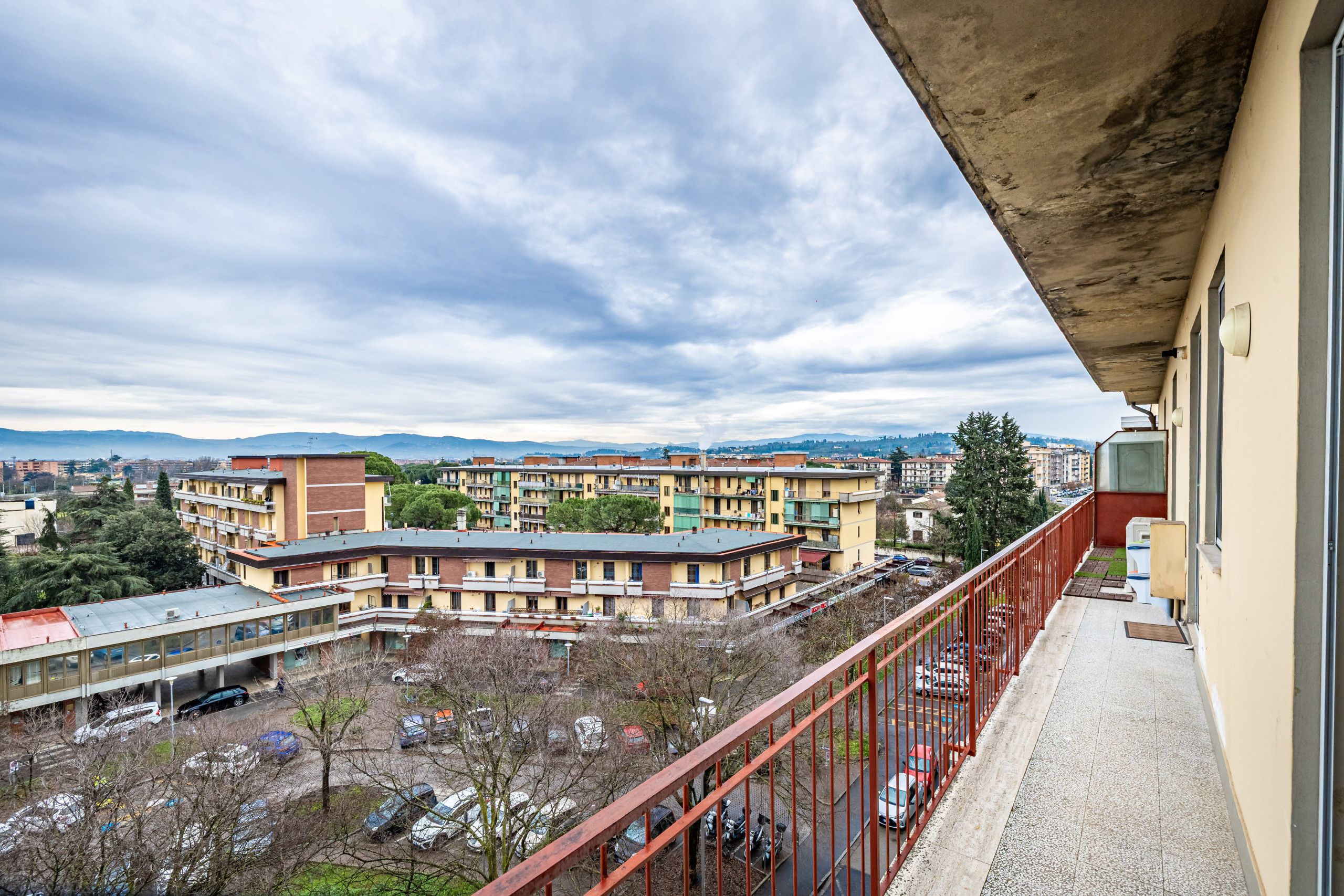 A balcony overlooking a parking area with multiple buildings in the background.
