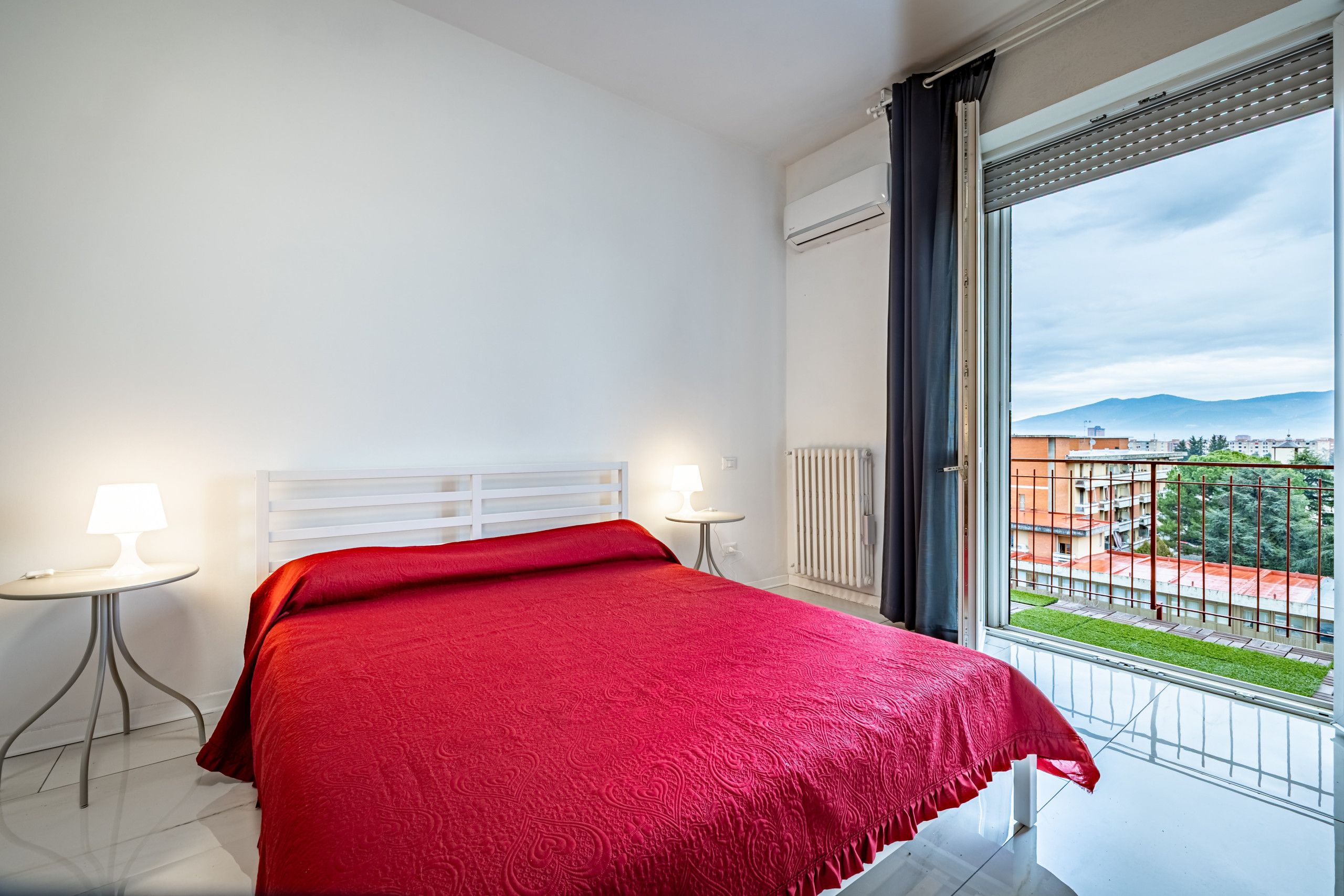 Et soveværelse med en seng, to sengeborde med lamper, en radiator og et vindue med udsigt til bygninger og grøntområder.