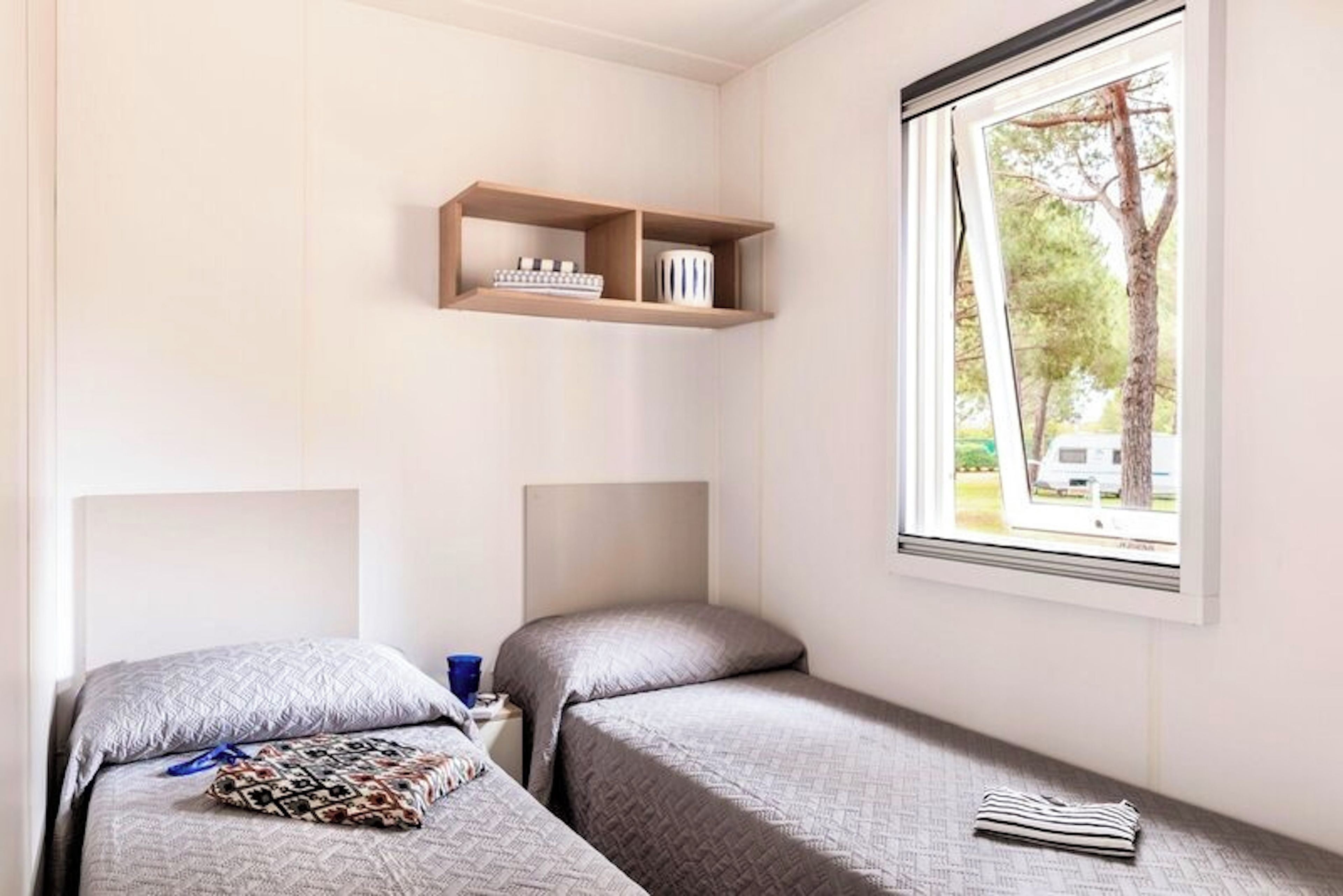 Een slaapkamer met twee eenpersoonsbedden, een plank boven de bedden en een raam met uitzicht op bomen en een voertuig buiten.
