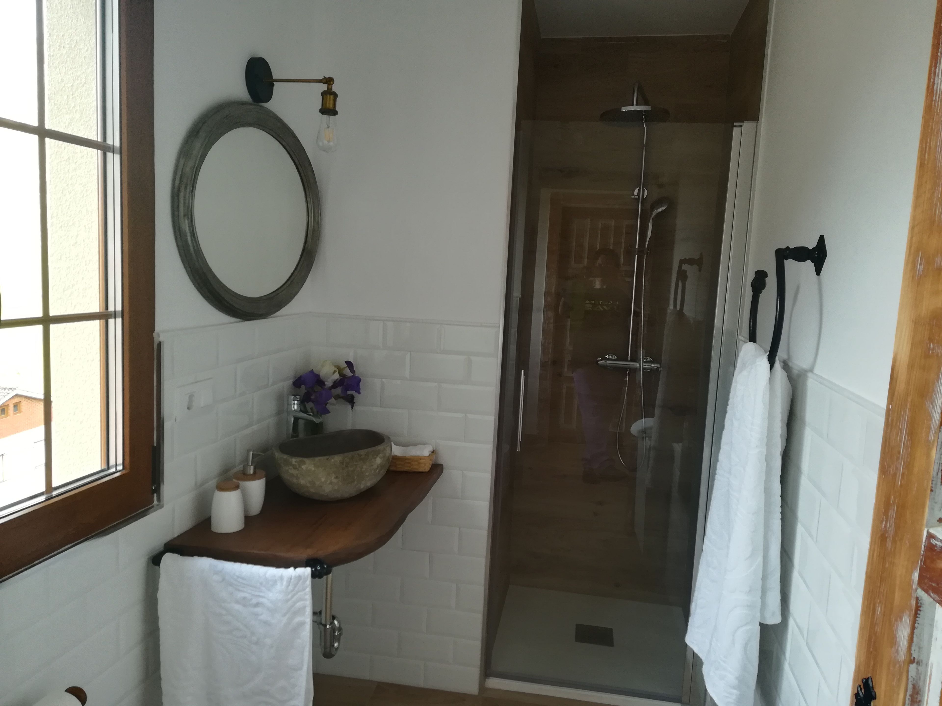 Een badkamer met een houten wastafelmeubel, een ronde spiegel, een douche met glazen wanden en een raam.