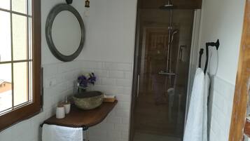 Een badkamer met een houten wastafelmeubel, een ronde spiegel, een douche met glazen wanden en een raam.