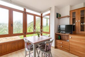 Une pièce comprenant une table à manger en bois, deux chaises, un téléviseur posé sur un meuble en bois et une grande fenêtre donnant sur un paysage verdoyant.