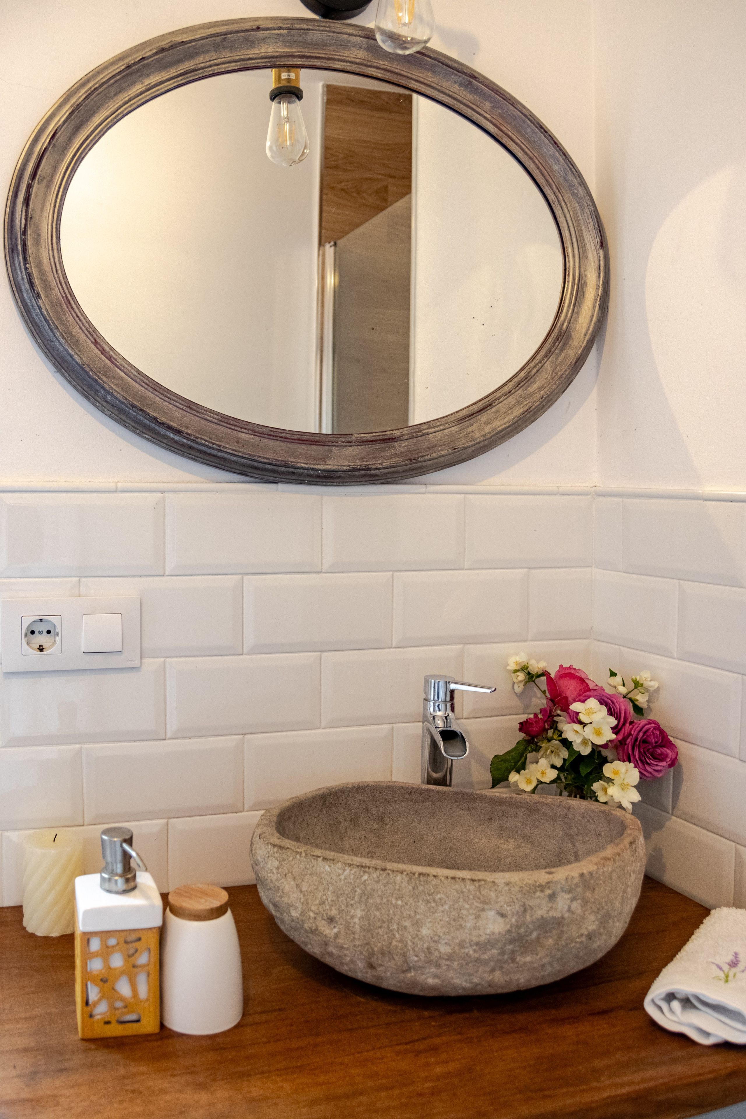 Une salle de bain avec un miroir rond, un lavabo en pierre, un robinet, un luminaire et un vase rempli de fleurs.