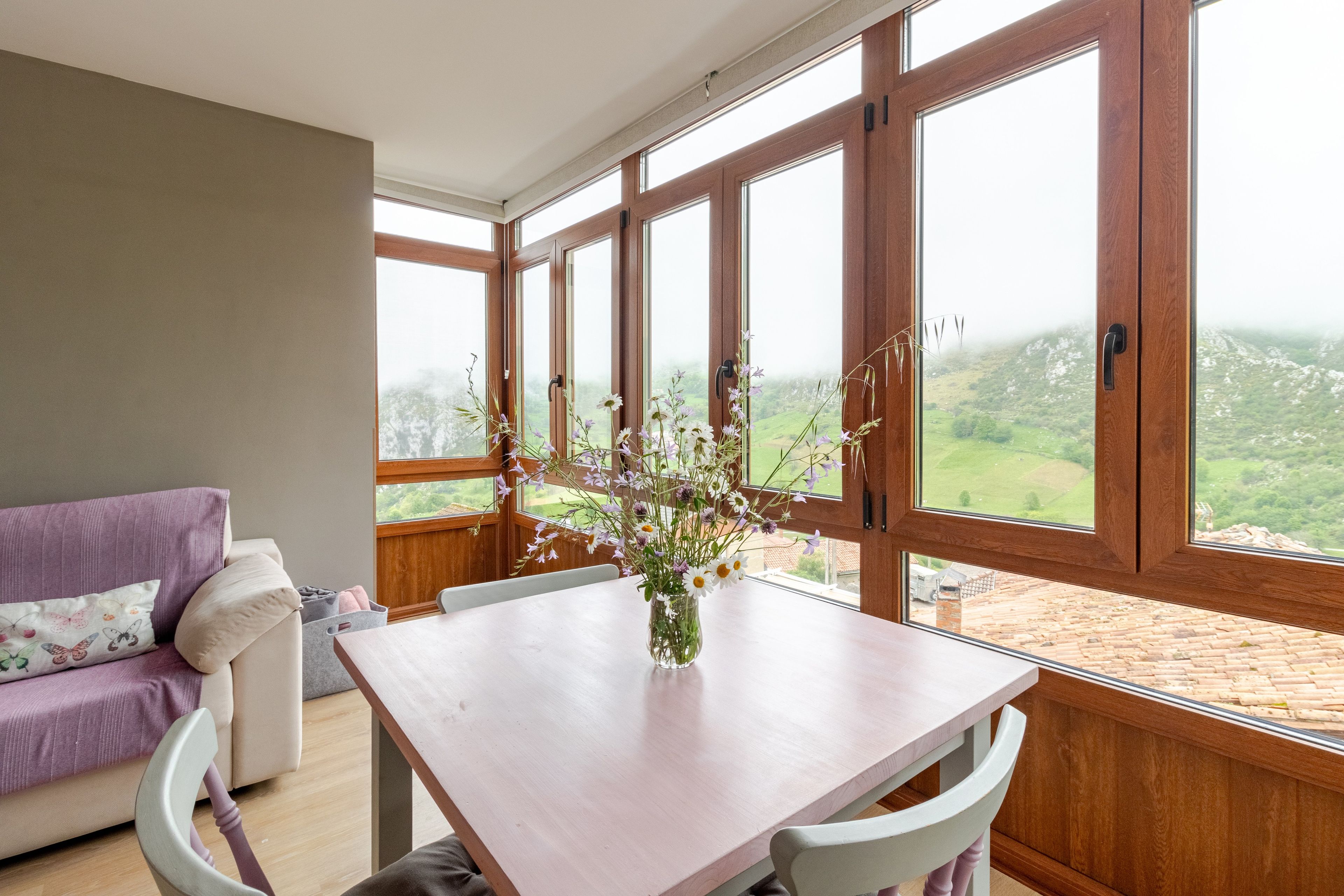 Een kamer met een houten tafel en stoelen, een paarse bank en een groot raam met uitzicht op een groen landschap.