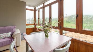Een kamer met een houten tafel en stoelen, een paarse bank en een groot raam met uitzicht op een groen landschap.
