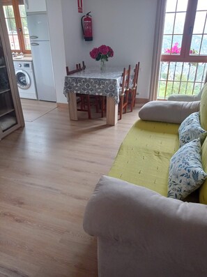 A living room with a sofa, a dining table with chairs, a fire extinguisher on the wall, and a window with a view.