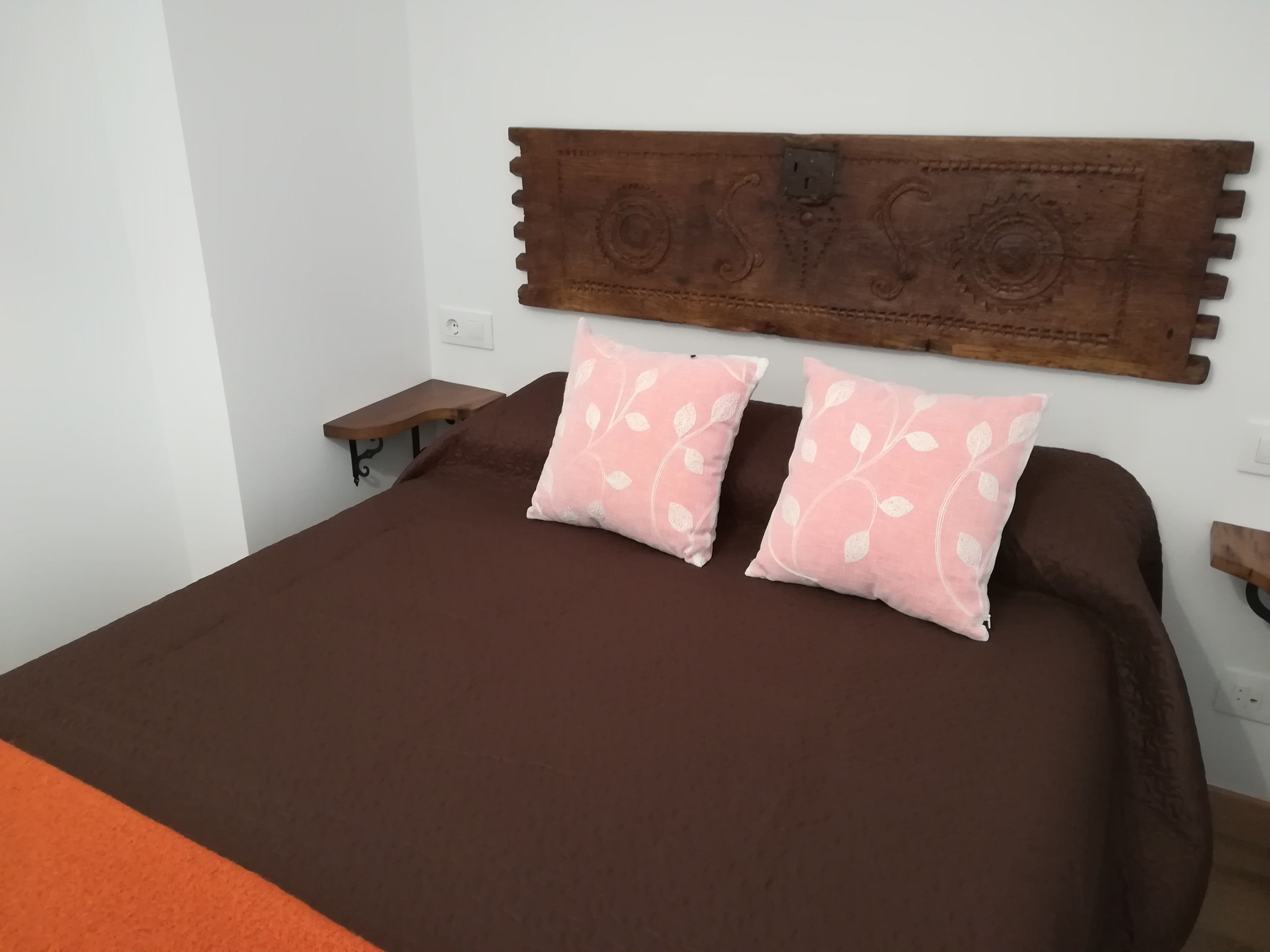 Una cama con cabecera de madera, dos almohadas rosas con diseños de hojas y una colcha marrón.
