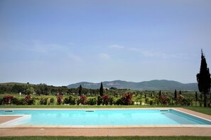 Une piscine rectangulaire à l’eau d’un bleu limpide, entourée d’une zone pavée. Au fond, on aperçoit un jardin bien entretenu, avec divers arbustes et arbres, et une vue sur des collines lointaines.