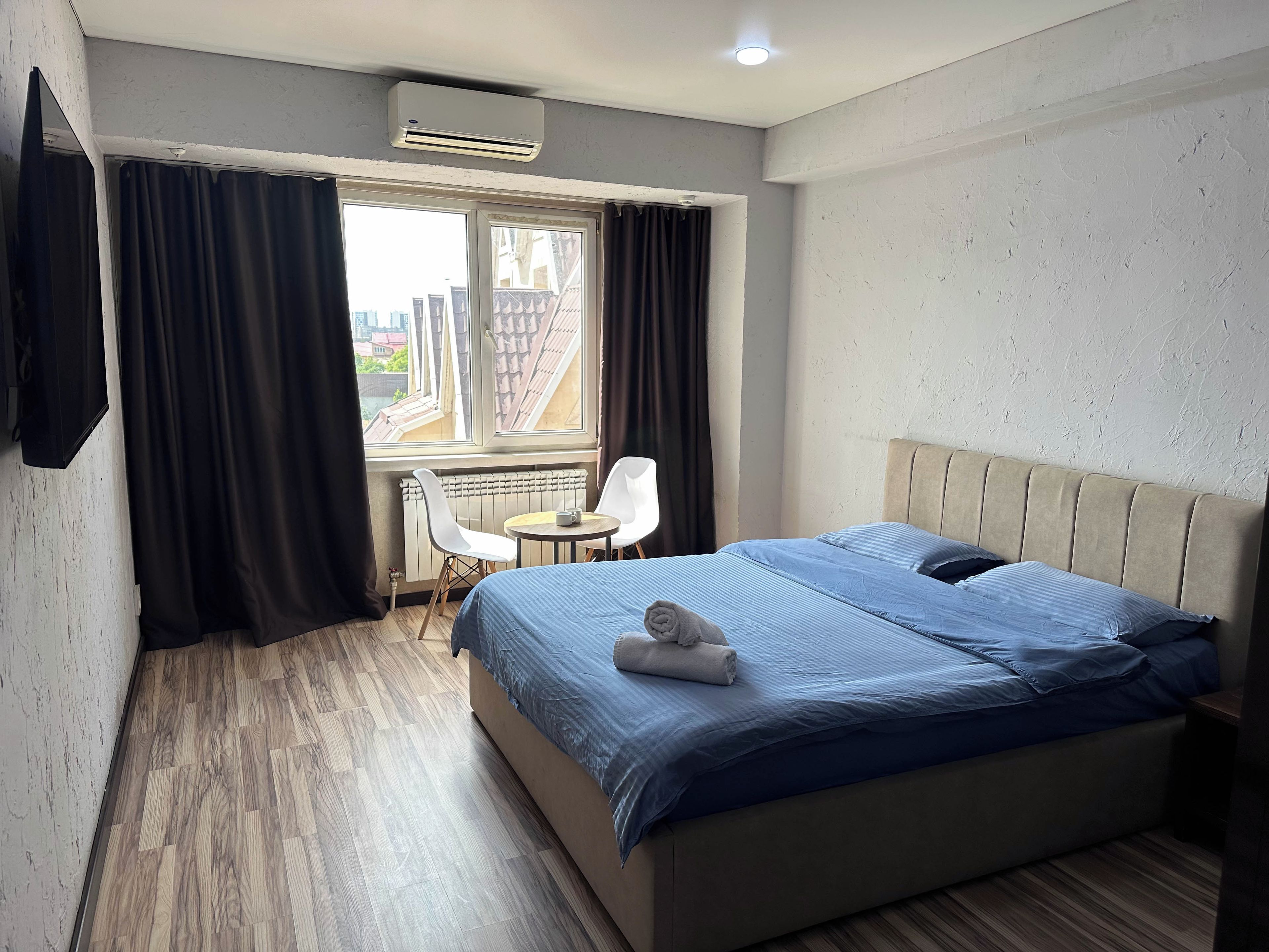 A hotel room with a bed, a small table, two chairs, a window with curtains, and an air conditioning unit.