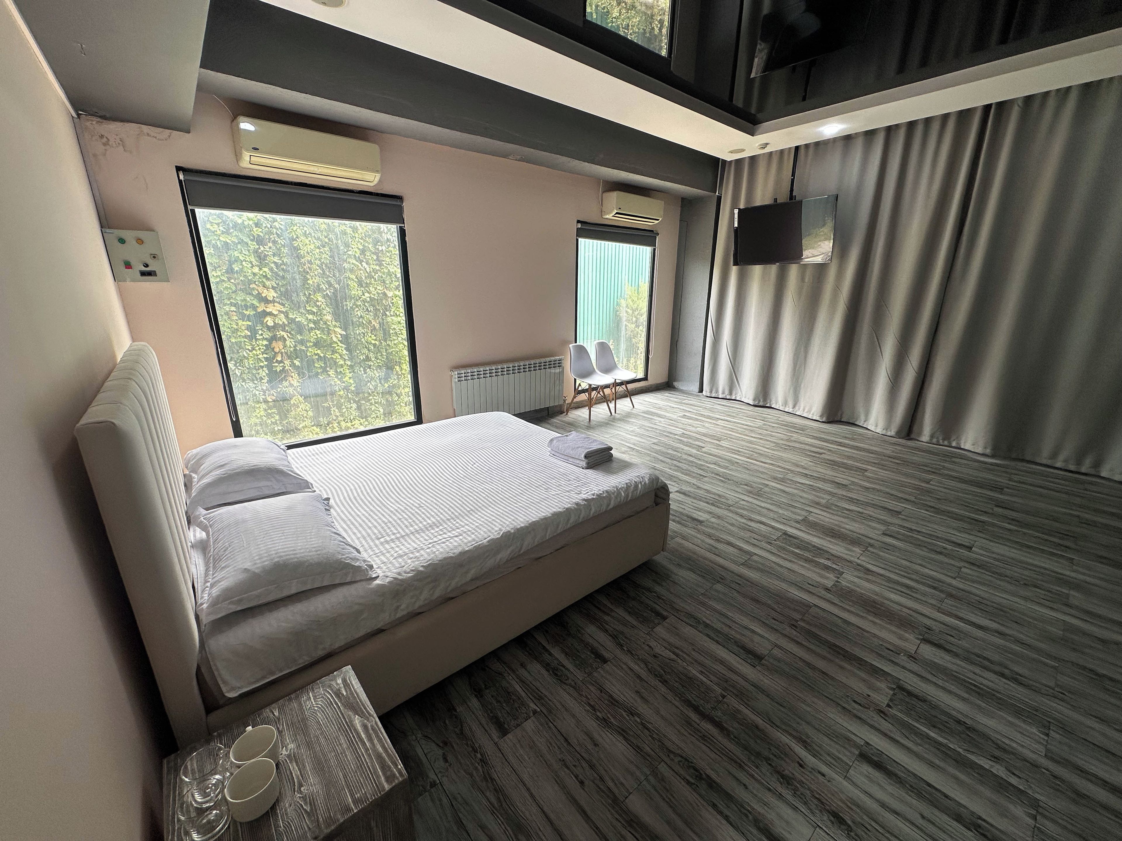 A modern hotel room with a large bed, a window with a view of trees, a radiator, and a flat-screen TV mounted on the wall.