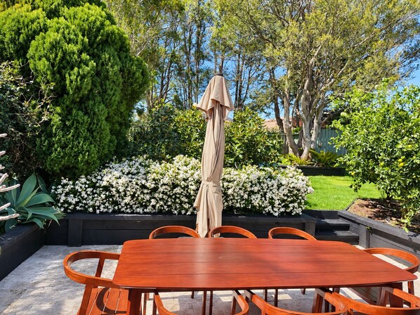 Un espace de restauration en plein air avec une table et des chaises en bois, entouré d’une végétation luxuriante et de plantes en fleurs.
