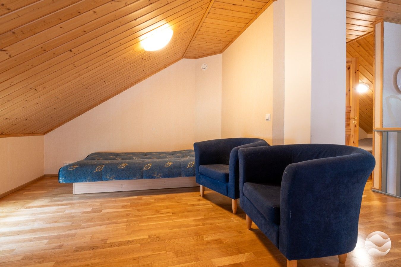 Quarto em estilo loft com teto inclinado, cama com colcha estampada, duas poltronas e piso de madeira.