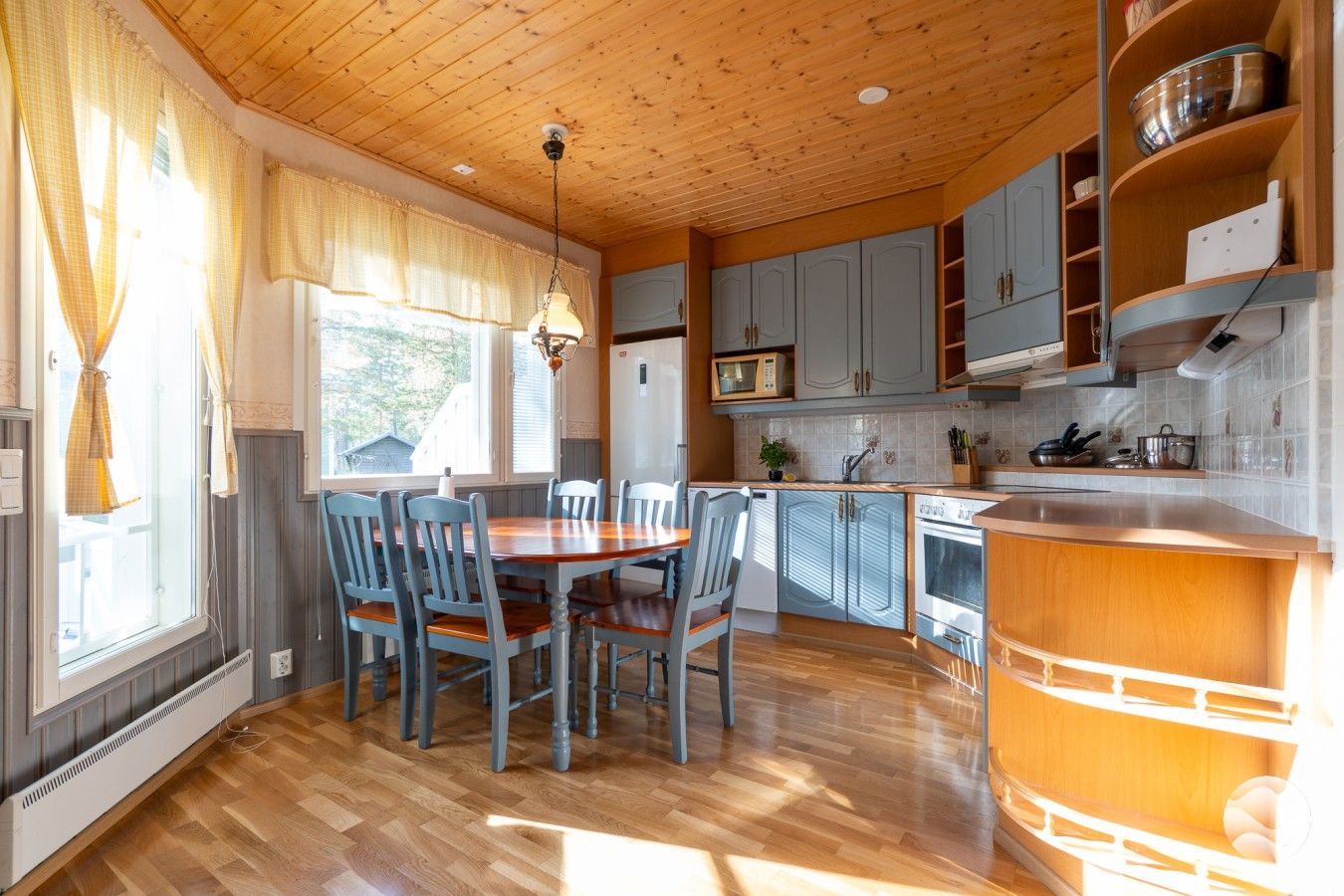 Una cocina con armarios de madera, una mesa de comedor con sillas y un ventanal con cortinas.