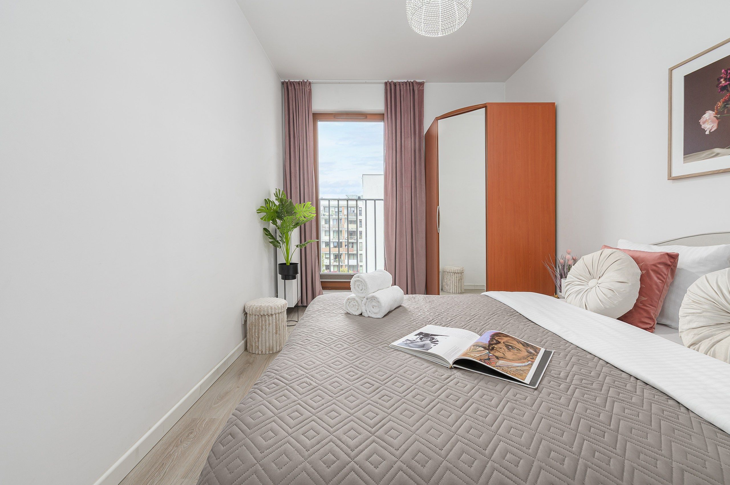 A modern hotel room with a bed, a window with curtains, a potted plant, and a magazine on the bed.