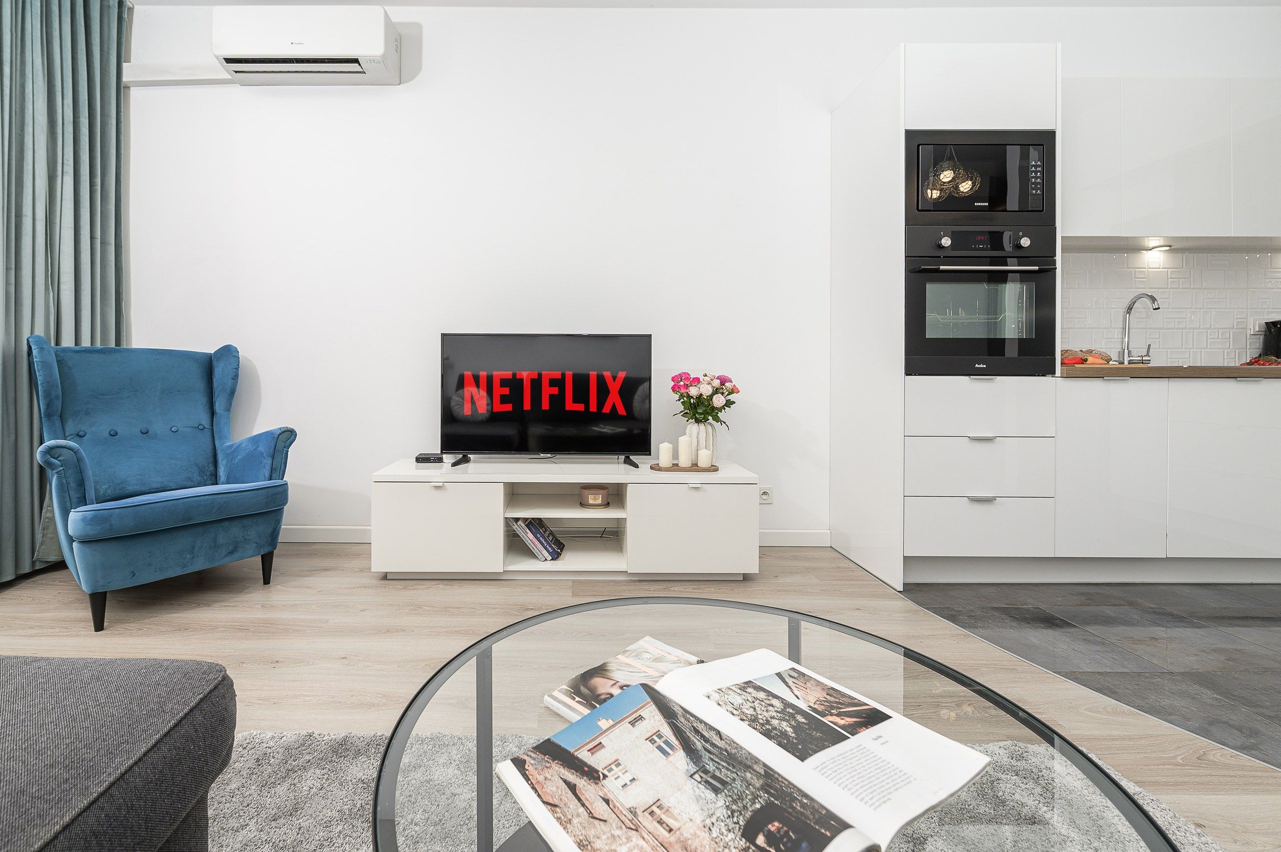 A modern living room with a blue sofa, a flat-screen TV, a glass coffee table, and a kitchen area with white cabinets and appliances.