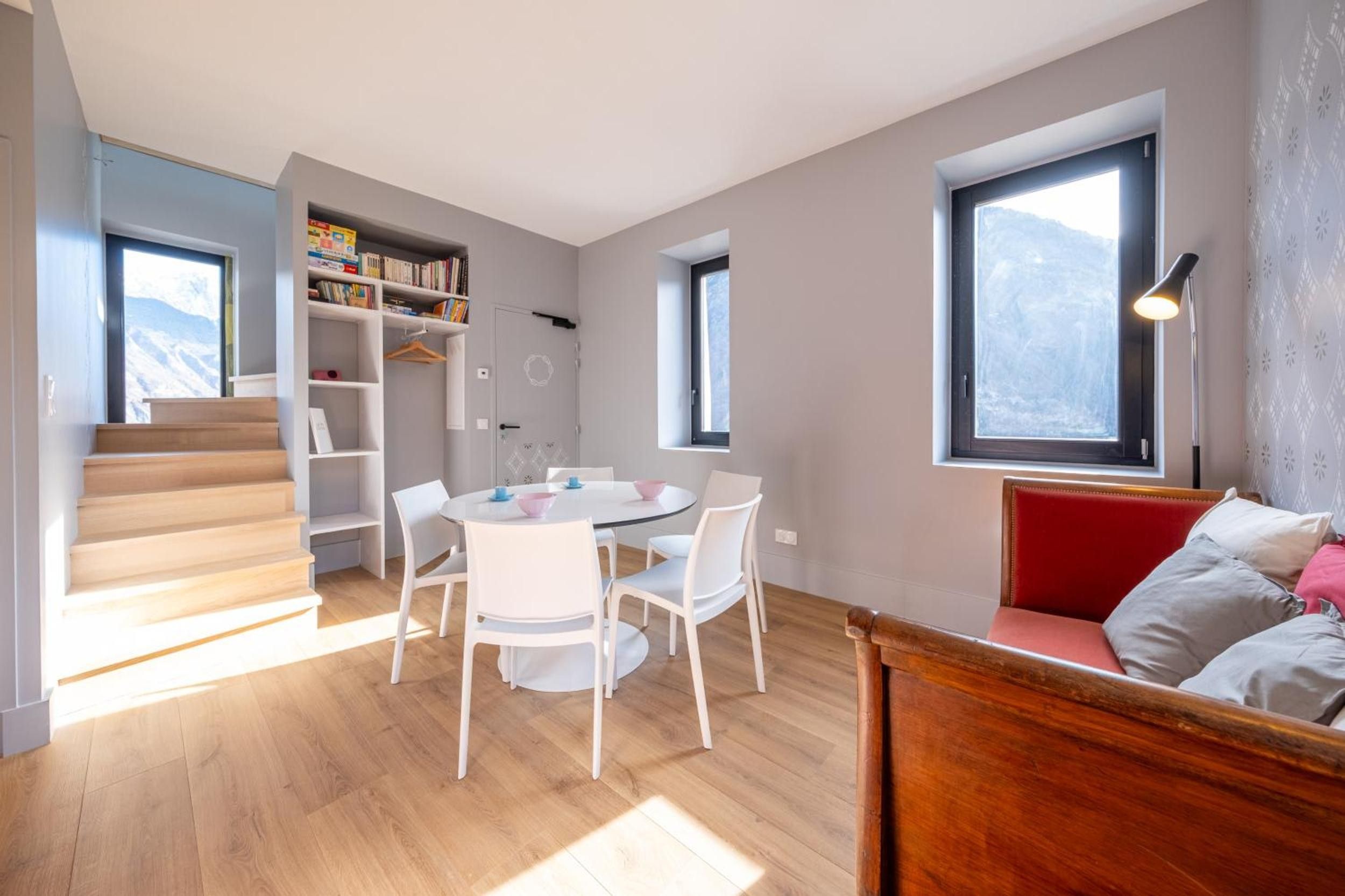 Un intérieur moderne avec une table de salle à manger ronde, des chaises blanches, une bibliothèque, un escalier et un canapé rouge.
