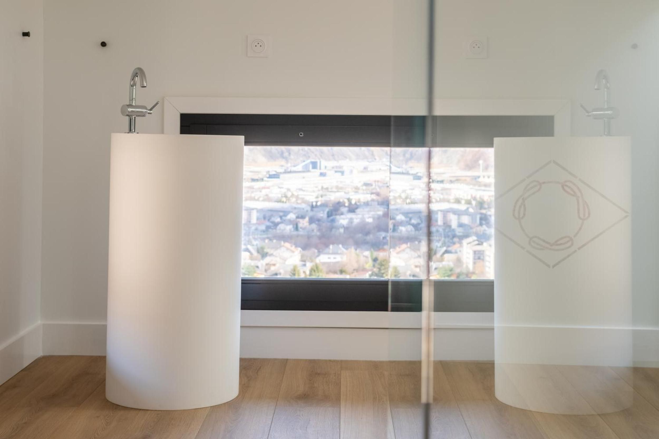 Ein Zimmer mit zwei weißen, zylindrischen Säulen, einem Fenster mit Blick auf eine Stadtlandschaft und einem Holzboden.