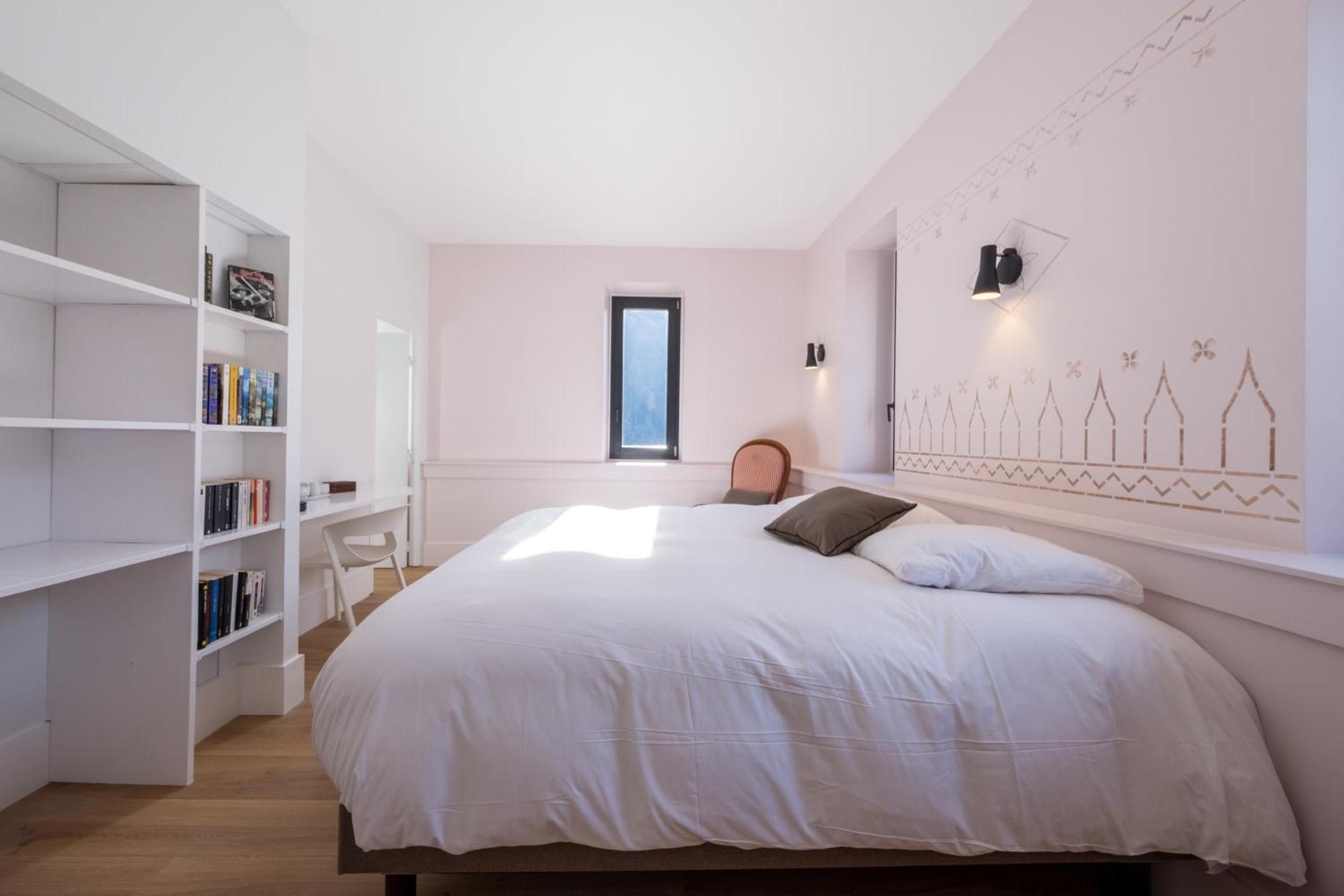 Een slaapkamer met een bed, een boekenkast, een stoel en een muur met een geometrisch patroon.