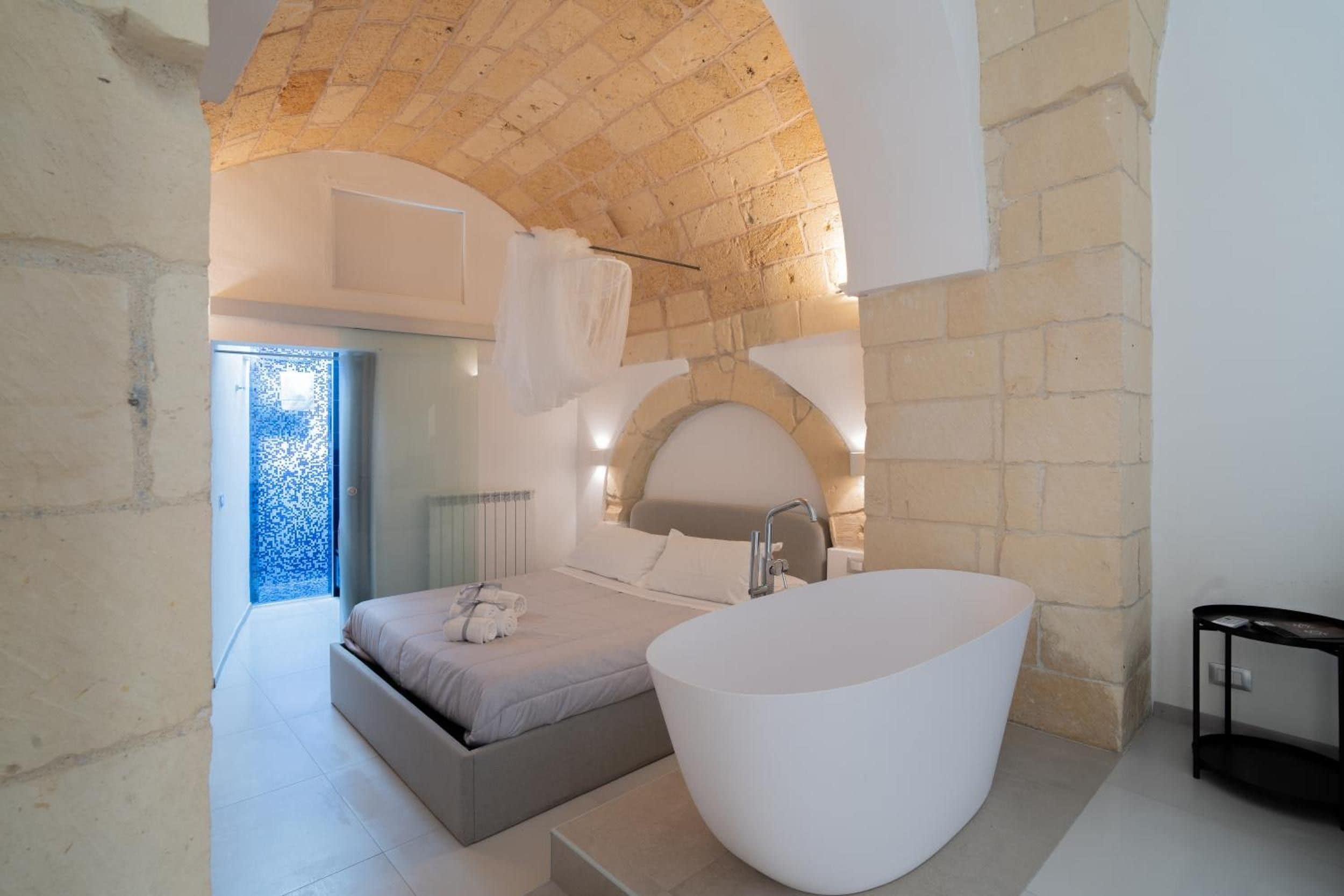 A modern bedroom with a large bed, a freestanding bathtub, and a stone wall.