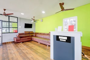 A room with a sectional sofa, a ceiling fan, a television mounted on the wall, and a reception desk with a sign. - Palette RC Resort  Kovalam Beach ECR (Chennai)