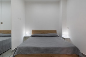 Ein Einzelbett mit Holz Kopfteil und einer grauen gesteppten Bettdecke in einem Zimmer mit weißen Wänden und einem kleinen Nachttisch mit Lampe.
