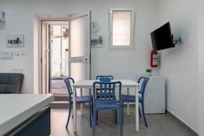 Une pièce avec une table blanche et des chaises bleues, un petit appareil électroménager blanc, un téléviseur fixé au mur et un extincteur sur le mur.