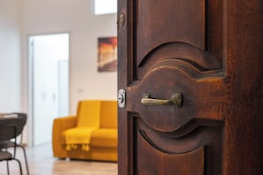 Uma porta de madeira com maçaneta de latão e fechadura, dando acesso a um quarto com sofá amarelo e piso de madeira.