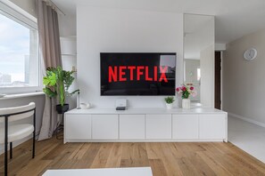 Un salon moderne avec un grand téléviseur à écran plat fixé sur un mur blanc, un meuble de rangement blanc en dessous, et une vue sur la ville à travers une fenêtre ornée de rideaux.