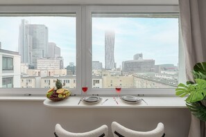 A dining area with a table set for two, a fruit bowl, and two glasses of red wine on a windowsill with a cityscape view. - Lumina 3 room Apart near Pałac Kultury (Warszawa)
