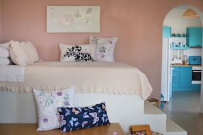 A bedroom with a bed, pillows, a bedside table, and a view into a kitchen area. - Elefthia Syros-Luxury Houses & Suites (Syros)
