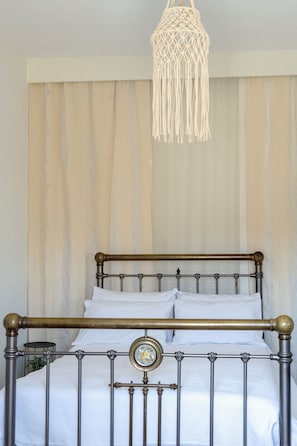 A bed with a metal frame and a decorative headboard, a macrame hanging light fixture, and a curtain with a striped pattern. - Elefthia Syros-Luxury Houses & Suites (Syros)