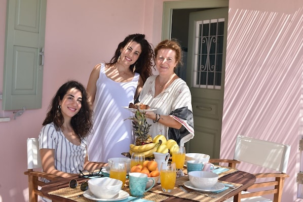 A table set with breakfast items, including fruits and beverages, in a room with a pink wall and a window with bars. - Elefthia Syros-Luxury Houses & Suites (Syros)