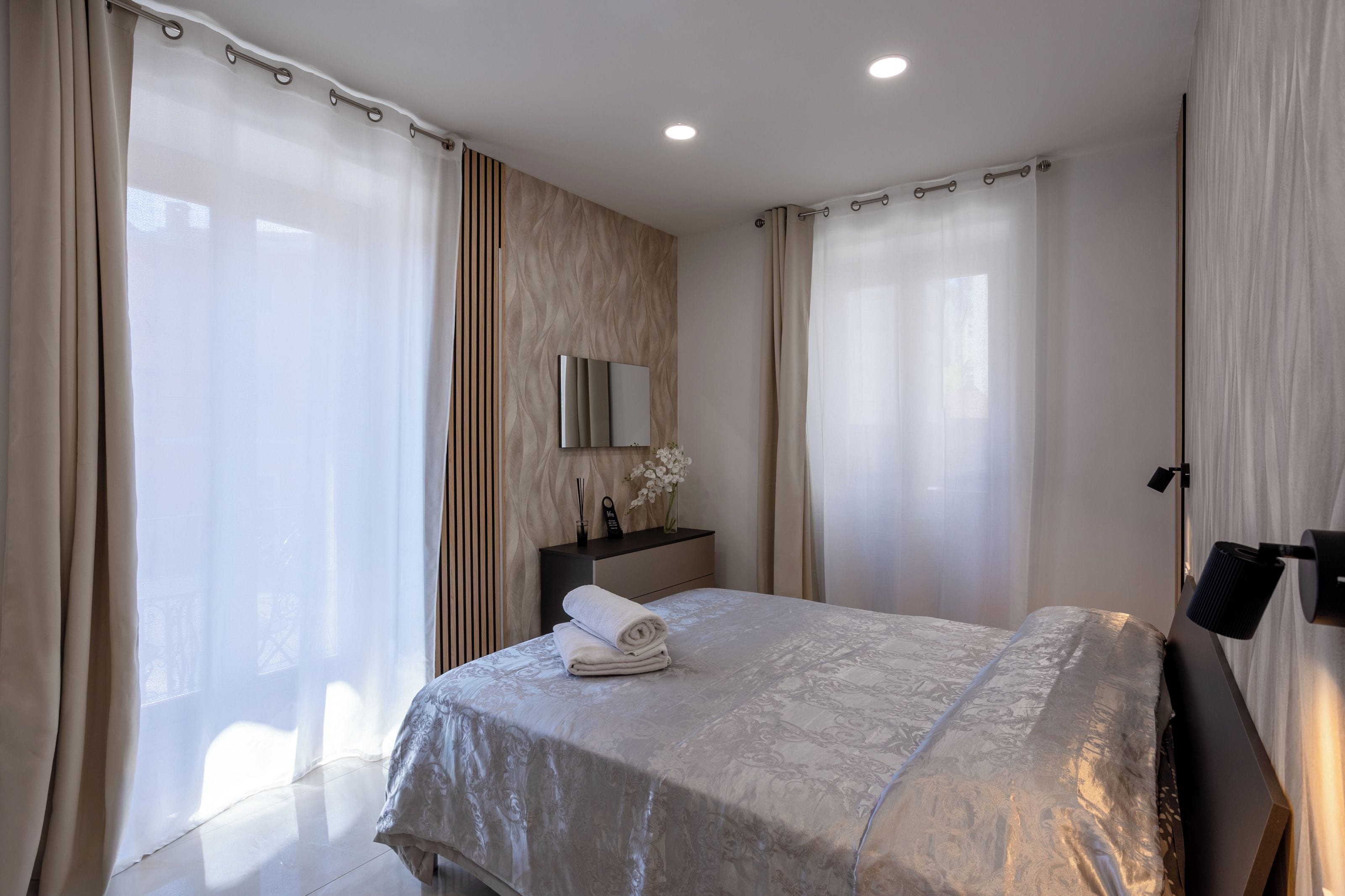A hotel room with a bed, a bedside table with a lamp, a mirror, and curtains.