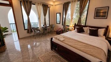 A hotel room with a large bed, a sitting area with a chair, a small table, and a view of the outdoors.