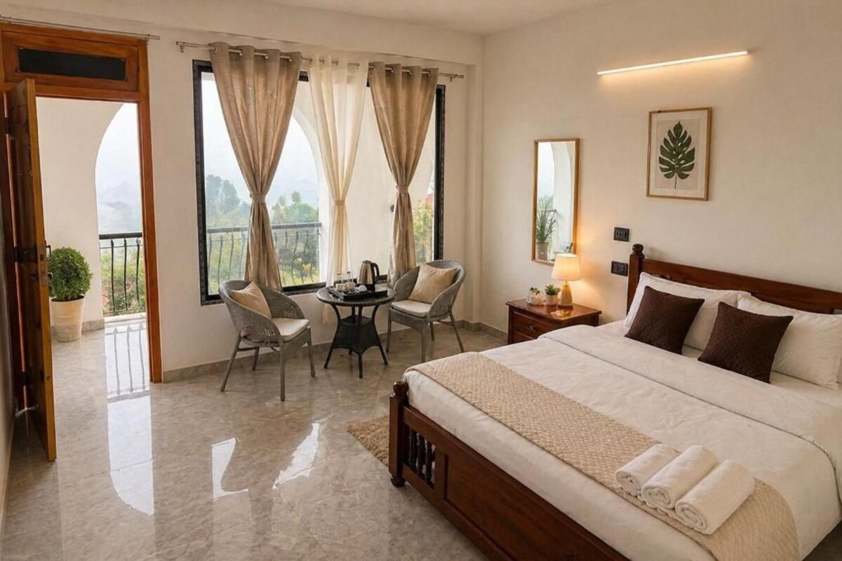 Ein Hotelzimmer mit Bett, Schreibtisch, zwei Stühlen, einem Balkon mit Vorhängen und Blick ins Freie.