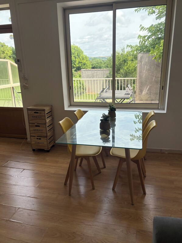 A dining area with a glass table and yellow chairs, a wooden crate labeled 'STORAGE', and a view of a balcony with a railing.