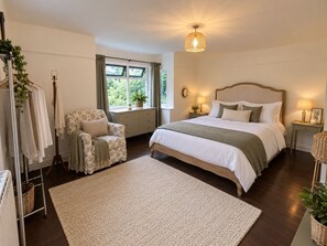 Ein Schlafzimmer mit einem Bett, einem Sessel, einer Kommode, einem Fenster mit Vorhängen, einer Pendelleuchte und einer Heizung.
