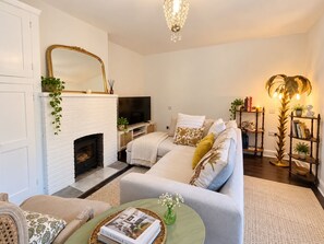 Ein Wohnzimmer mit einem weißen Kamin, einem Fernseher, einer Couch mit Zierkissen, einem Couchtisch mit einem Buch und einem Bücherregal mit Büchern und Dekorationsobjekten.