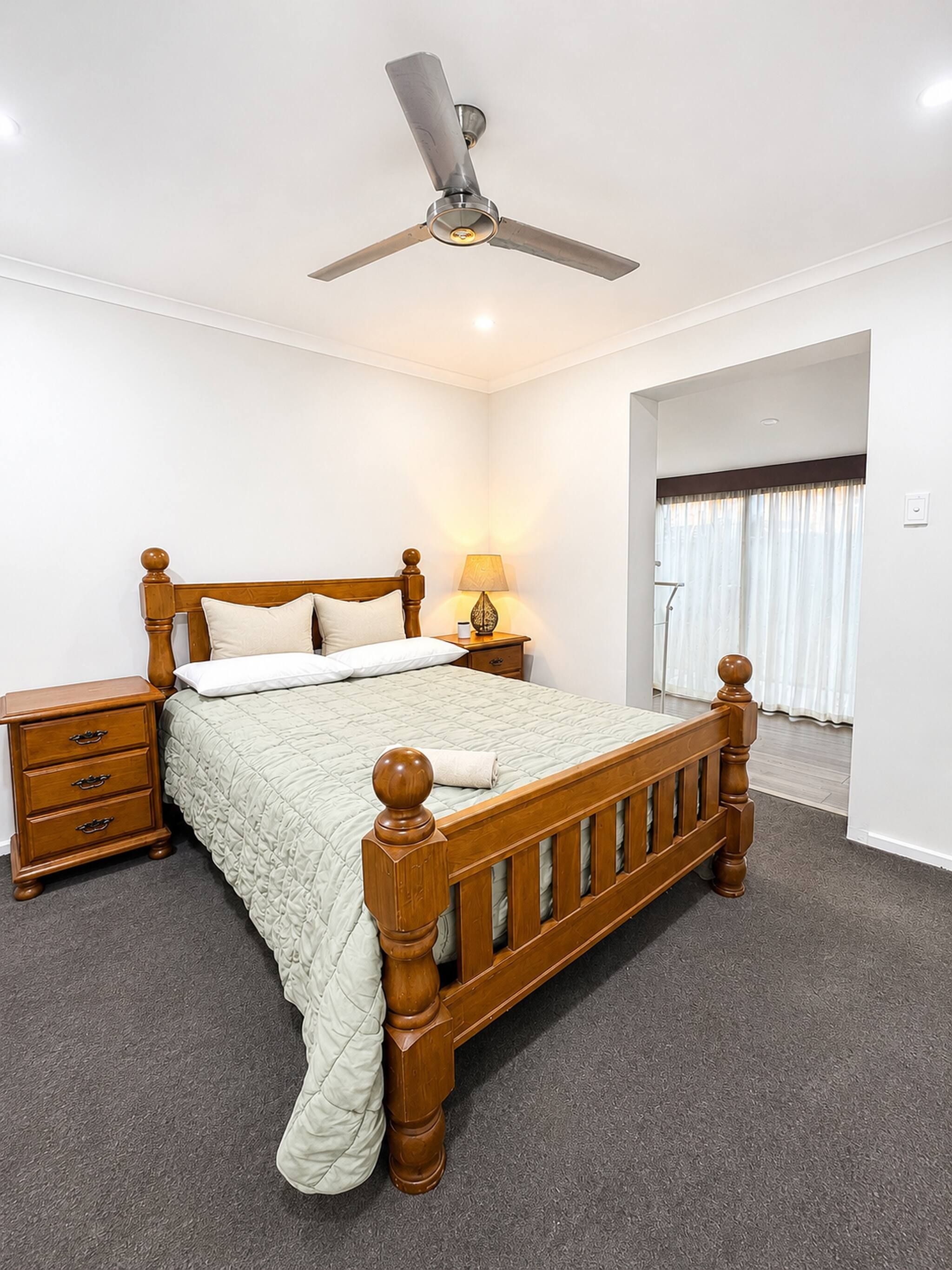 A bedroom with a wooden bed, a nightstand, a lamp, a ceiling fan, and a window with curtains.