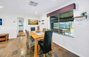 A dining area with a wooden table and chairs, a kitchen in the background, and a large window with a view of the outdoors. - Gates Mini Ranch Waterloo Corner (Waterloo Corner)