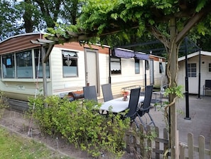 A mobile home with a covered patio area, a table with chairs, and a small garden. - Gezinsvakantie in de Drentse natuur | Stacaravan (Wijster)