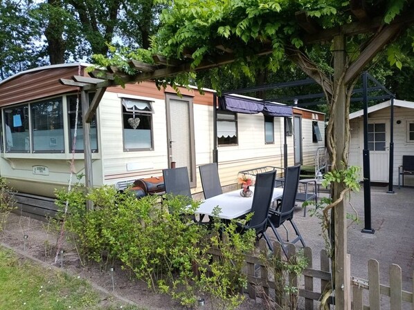 A mobile home with a covered patio area, a table with chairs, and a small garden. - Gezinsvakantie in de Drentse natuur | Stacaravan (Wijster)