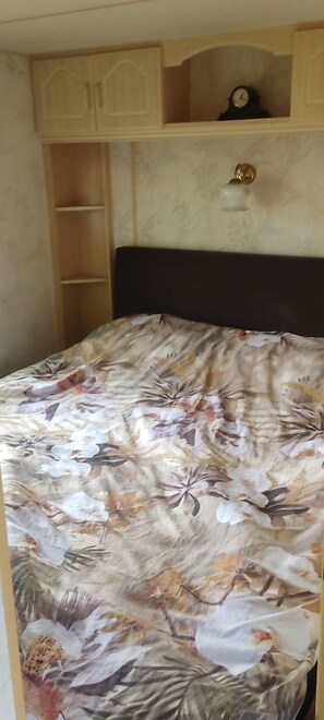 A bed with a floral bedspread, a wooden headboard, and a clock above it. - Gezinsvakantie in de Drentse natuur | Stacaravan (Wijster)