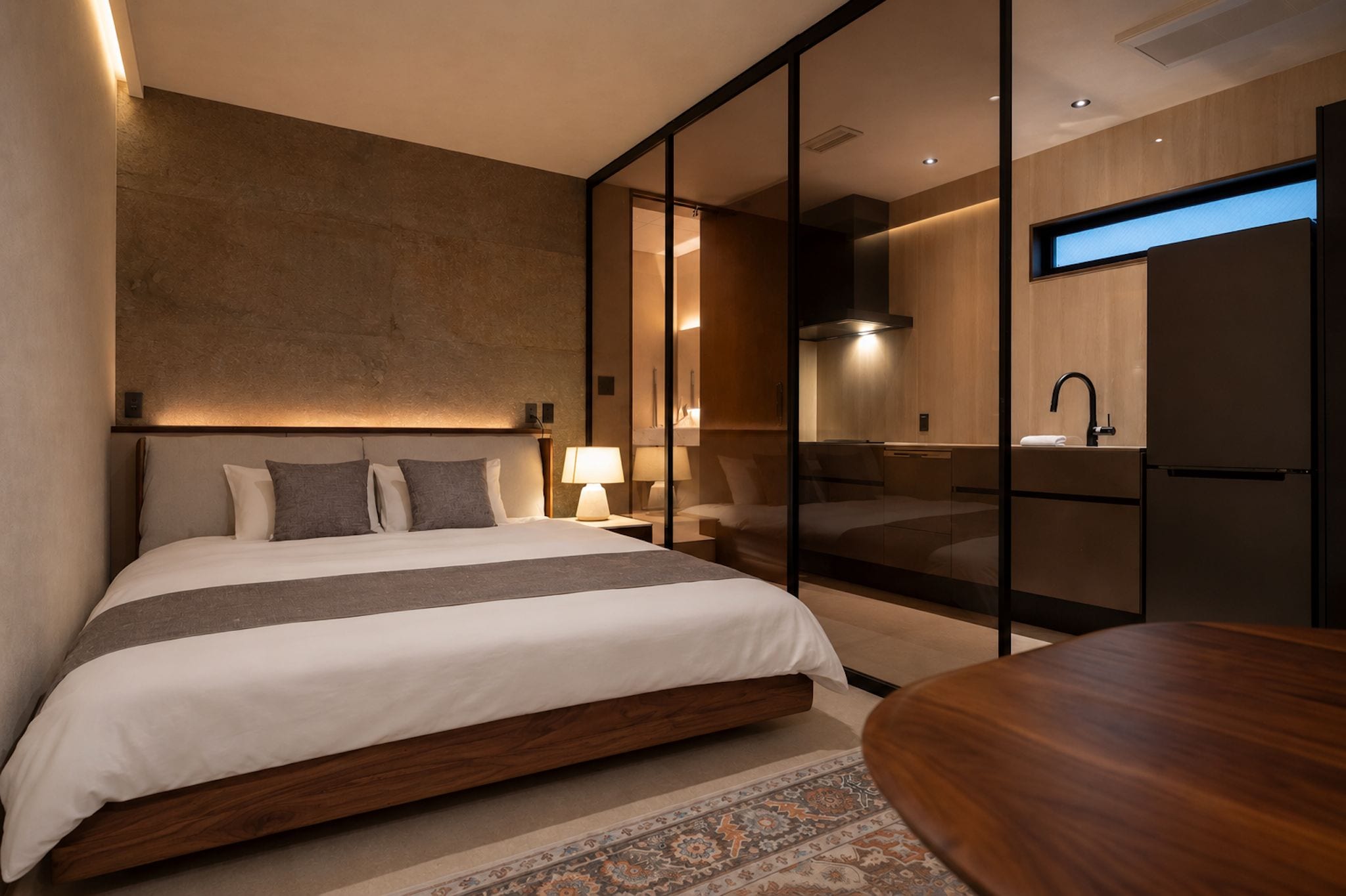 Een moderne hotelkamer met een groot bed, een houten nachtkastje, een inbouw bureau en een glazen wand die de slaapkamer van de badkamer scheidt.