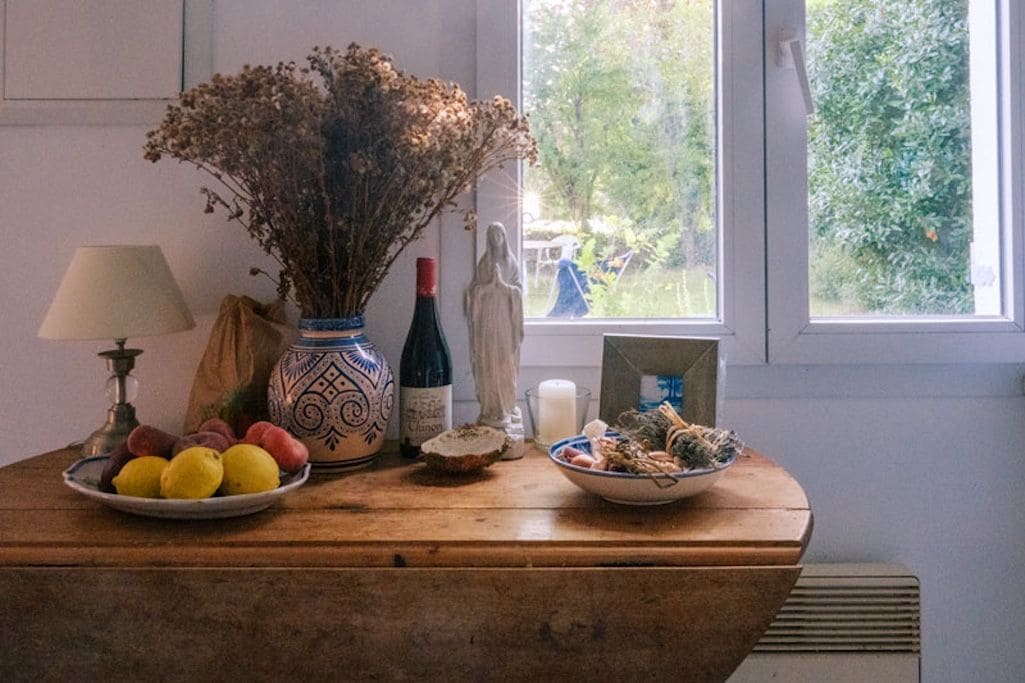 A wooden table with a lamp, a vase with dried flowers, a bottle, a small statue, and a bowl of food.